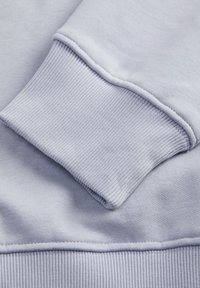 Jasnoniebieska bluza z tkaniny z prążkowanym mankietem. Gładka tekstura i widoczne przeszycia wzdłuż szwu, podkreślające prosty design.