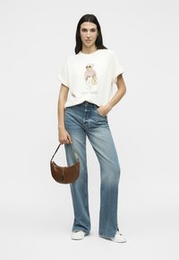 Γυναίκα που φοράει λευκό γραφικό t-shirt, τζιν με ψηλή μέση και φαρδιά μπατζάκια, λευκά αθλητικά παπούτσια, κρατώντας καφέ τσάντα σε λευκό φόντο.