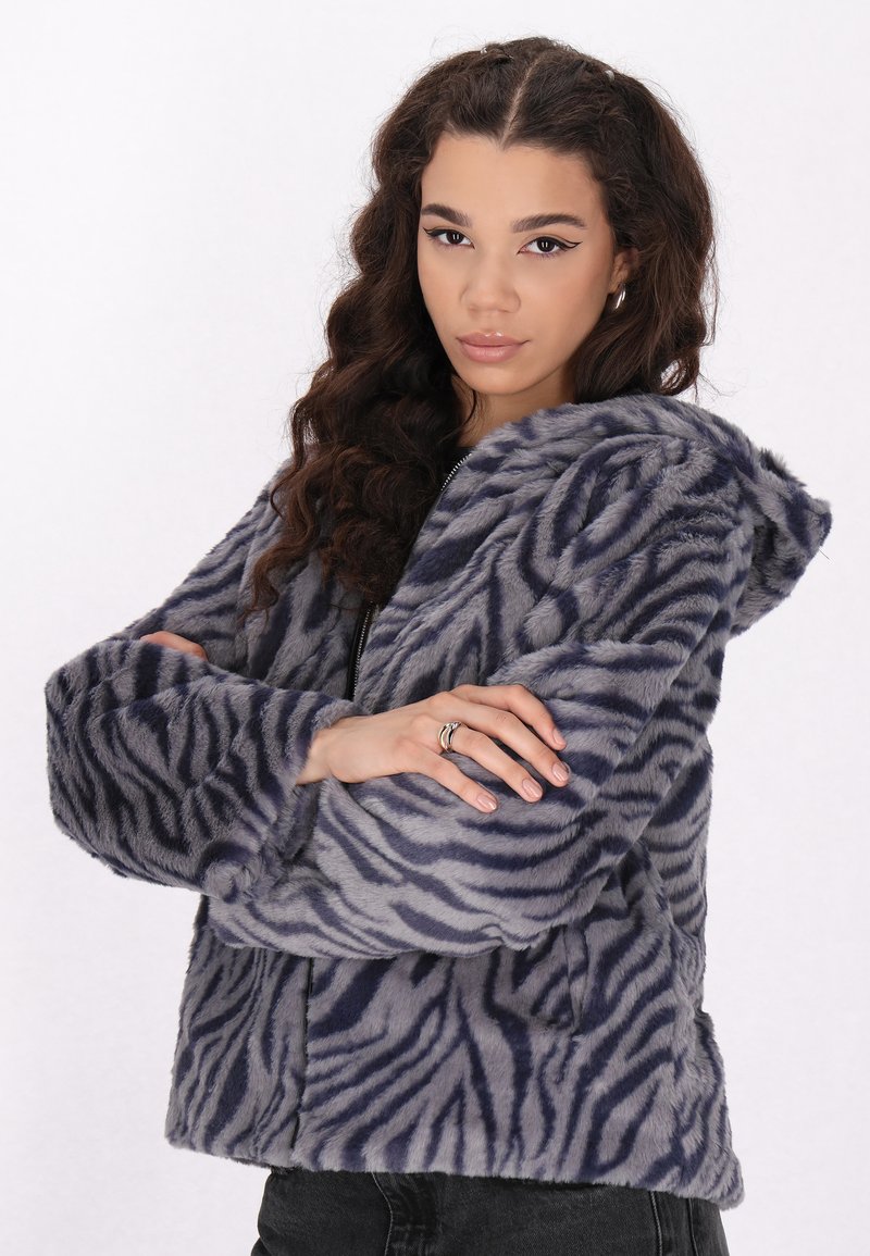 Jachetă cu glugă din blană artificială, de culoare gri, cu dungi în stil zebra bleumarin. Previne închidere cu fermoar, mâneci late și buzunare laterale. Finisaj moale, texturat.