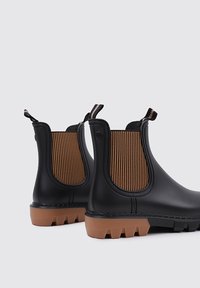 Bottes Chelsea en caoutchouc noir avec des panneaux élastiques sur les côtés, des rayures horizontales brunes et une semelle brune texturée ; languettes pour un enfilage facile.
