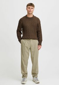 Brown knitted sweater with a cable pattern, paired with high-waisted beige trousers and multicolored athletic shoes.