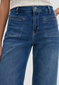 Donkerblauwe denim jeans met een hoge taille, vijf zakken en een subtiele vervaagde structuur. Bevat een knoopsluiting en stikseldetails.