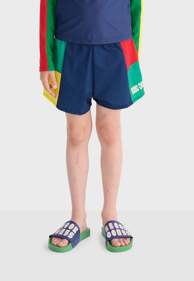 Child wearing navy blue shorts, multicolor long-sleeve shirt, and blue-green slide sandals with white text on straps standing on light background.
