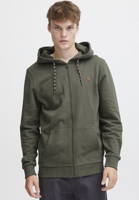 Haut à capuche zippé vert olive en tissu doux, avec une capuche à cordon de serrage, des poches avant et un petit logo brodé sur la poitrine.