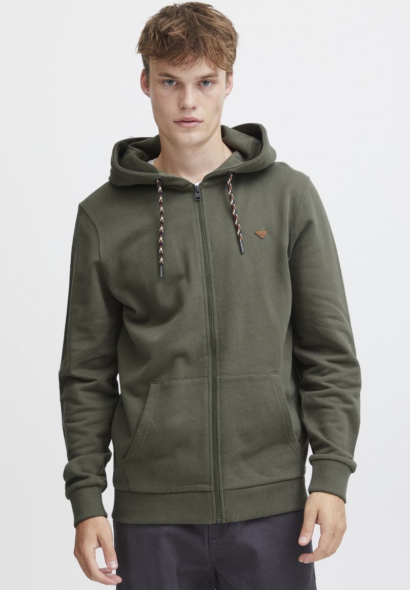 Haut à capuche zippé vert olive en tissu doux, avec une capuche à cordon de serrage, des poches avant et un petit logo brodé sur la poitrine.