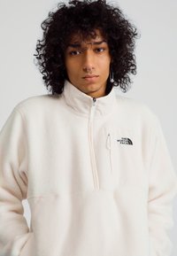 Pullover in pile bianco con colletto alto, zip a quarto e tasca sul petto con logo nero. Texture morbida e vestibilità rilassata.