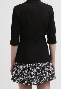 Blazer noir cintré avec manches trois-quarts, associé à une jupe à imprimé floral noir texturé avec des accents blancs et roses.