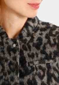 Giacca con stampa leopardata in tonalità di grigio e marrone, caratterizzata da un tessuto texture, colletto alto, chiusura con bottone e una piccola tasca sul petto.