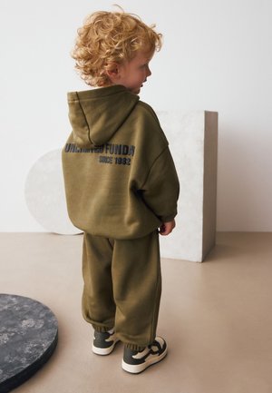 Olivengrøn hættetrøje og matchende bukser med en stor logo på ryggen. Blødt, struktureret stof og manchetter på ærmerne. Sneakers fuldender outfittet.