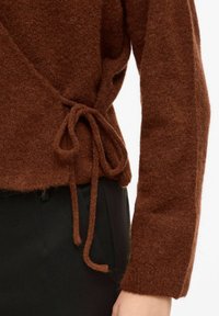 Brązowy wełniany sweter kopertowy z teksturowanym wykończeniem, z wiązaniem w talii i szerokimi rękawami. W zestawie z czarnymi spodniami.