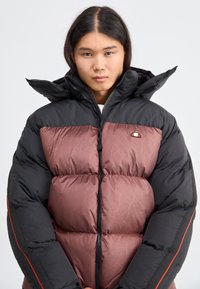 Chaqueta tipo puffer con la parte superior negra y la sección inferior acolchada en color burdeos con un patrón sutil. Tiene un cuello alto y un cierre frontal con cremallera.