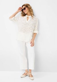 Weiße Augen-Spitze Bluse mit gebundenem Ausschnitt und aufgerollten Ärmel, kombiniert mit weißen cropped Hosen mit bestickten Säumen und beigen Slingback-Schuhen.