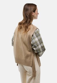 Gilet en tricot beige avec ourlet côtelé, superposé à une chemise à carreaux verte. Le gilet possède des nouettes sur les côtés et a une coupe décontractée.