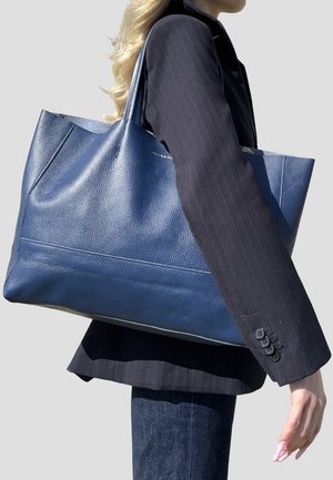 Donna con blazer scuro a righe sottili che porta una grande borsa tote in pelle blu texturizzata sulla spalla, su sfondo semplice