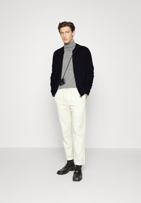 Black cardigan over a gray turtleneck, white trousers, and black shoes. Outfit features a crossbody bag and sleek design. Smooth textures.