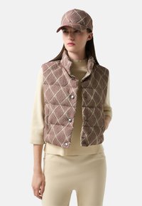 Padded vest z falistym wzorem w czerwono-kremowych odcieniach, z wysokim kołnierzem i zatrzaskami. Pasująca czapka w ten sam wzór dopełnia outfit.