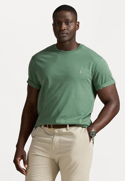 Grünes Baumwoll-T-Shirt mit umgeschlagenen Ärmeln und einem kleinen gestickten Logo. Kombiniert mit beigen Hosen und einer Uhr, die einen klassischen Schnitt und eine angenehme Textur aufweisen.
