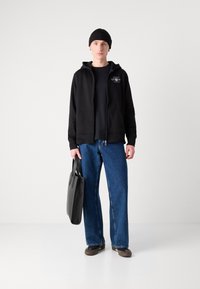 Crna zip-up trenirka preko crne majice, slobodne plave traperice, crna kapa, smešane cipele i siva torba. Jednostavan dizajn, ležeran stil.