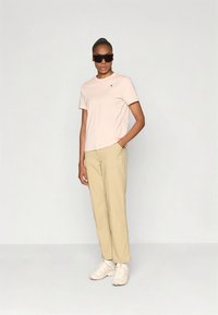 Svetloružové tričko s krátkym rukávom a modrým logom, v kombinácii s béžovými nohavicami a bielymi teniskami. Model nosí čierne slnečné okuliare.