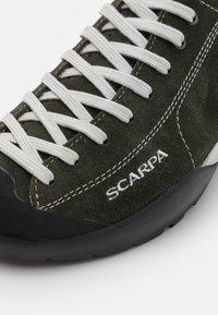 Grønn semsket klatresko med svart gummisåle, hvite lisser og "SCARPA"-logo. Har forsterket tå og kontrastsøm.
