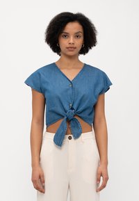 Top cropped de denim com decote em V, mangas curtas e soltas, e detalhe de laço na frente. Tecido azul com uma textura subtil. Fecho de botão no decote.