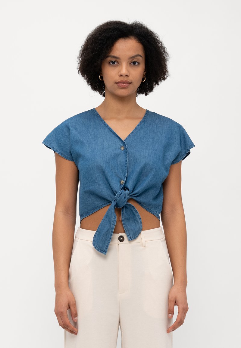 Top cropped de denim com decote em V, mangas curtas e soltas, e detalhe de laço na frente. Tecido azul com uma textura subtil. Fecho de botão no decote.