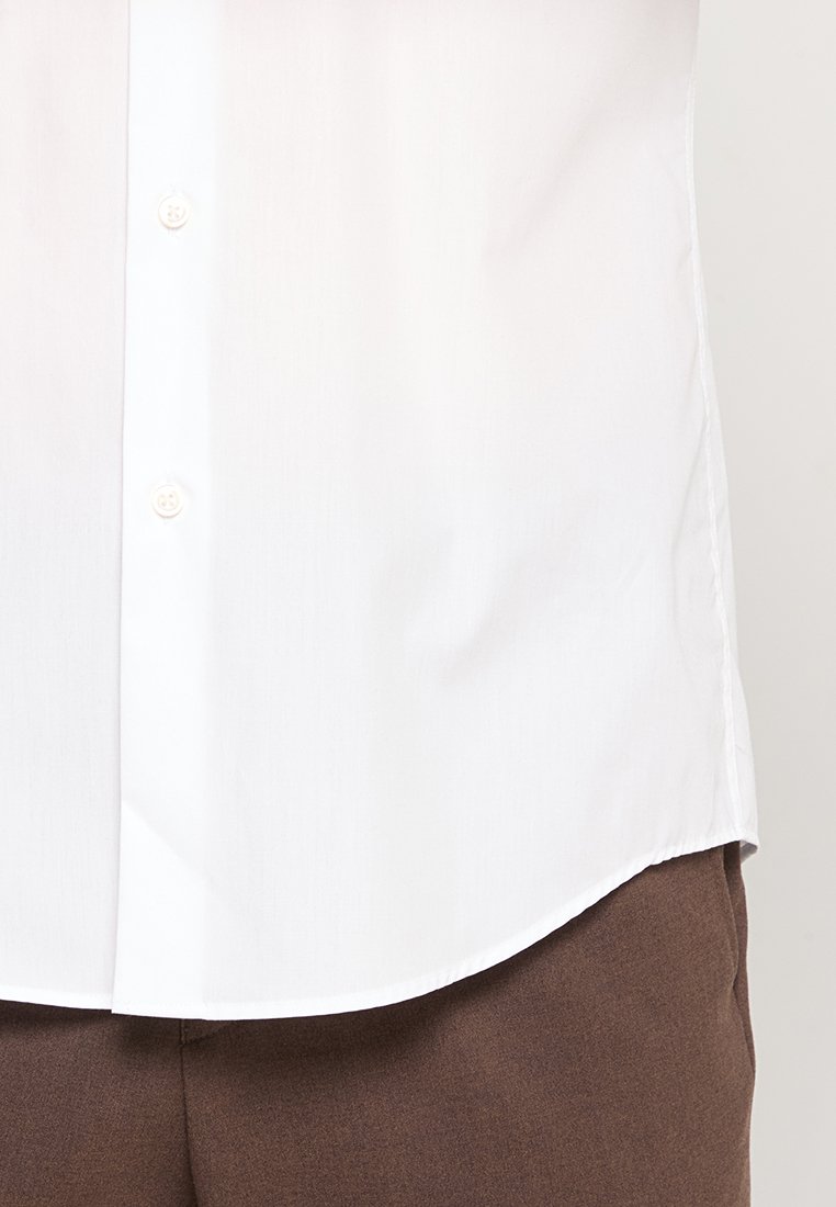 Camisa blanca de botones con una textura suave, dobladillo curvado y botones estándar, combinada con pantalones marrones.