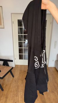 Mână care ține un hanorac negru GAP cu fermoar și litere albe, în interior, lângă o ușă cu geamuri și podea de lemn.