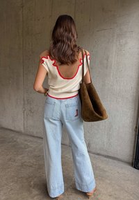 Γυναίκα με κυματιστά καστανά μαλλιά φοράει αμάνικο μπεζ και κόκκινο τοπ, ανοιχτό μπλε τζιν παντελόνι με φαρδύ πόδι και κέντημα με μπότα, και κρατάει μεγάλη καφέ τσάντα από σουέτ.