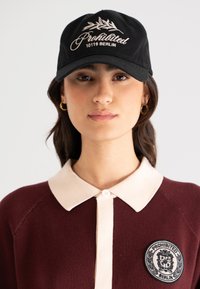 Svart baseballcaps med hvit broderi, som har "Forbudt" og "10119 Berlin." Mørk burgunder strikkegenser med beige krage og merke.