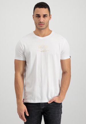Camiseta de algodón blanca con un cuello redondo, que presenta un diseño de logo dorado en el centro y una pequeña etiqueta negra en la manga izquierda.