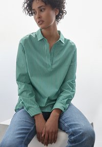 Zielona, w paski, długozaszerpowa koszula z kołnierzykiem i zapięciem, w połączeniu z jasnoniebieskimi dżinsami. Gładka faktura, luźny krój i boczne rozcięcia.