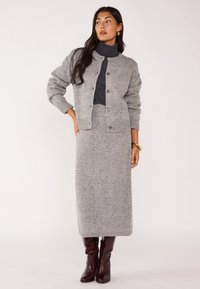Cardigan en tricot gris et jupe midi assortie, avec un motif texturé, fermeture à boutons et poignets côtelés. Associé à des bottes marron arrivant au genou.