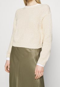 Lichtbeige, gebreide trui met een ronde halslijn en lange mouwen, gecombineerd met een gladde olijfgroene satijnen rok. Volledige weergave van beide kledingstukken.