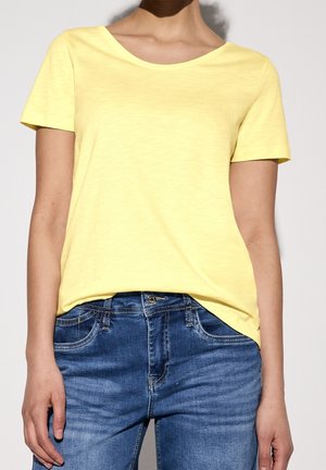 Vrouw draagt een effen geel T-shirt met korte mouwen, ingestopt in blauwe spijkerbroek, staand tegen een neutrale achtergrond.