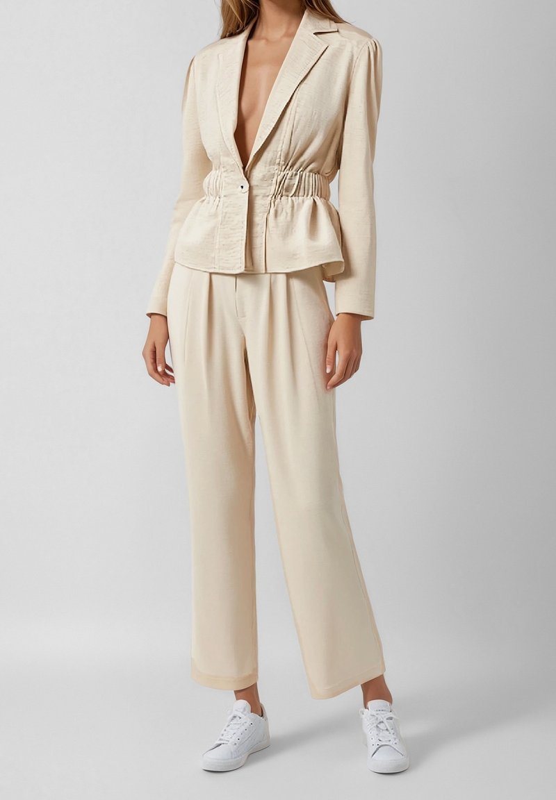 Femme portant un blazer beige froncé à la taille avec un pantalon large assorti et des baskets blanches sur fond uni.