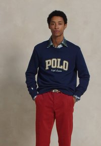 Marineblaues Sweatshirt mit "POLO"-Logo, über einem karierten Hemd getragen. Kombiniert mit roten Baumwollhosen und einem Gürtel. Schlichtes, lässiges Design.