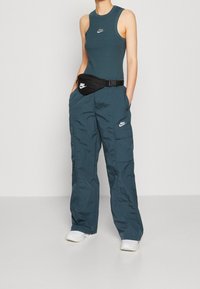 Person wearing teal sleeveless Nike top, matching cargo pants, white sneakers, and black Nike waist bag, standing against plain background.