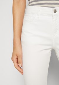 White jeans with a smooth texture, five-pocket design, and a metal button at the waist. The fabric is denim and stretches slightly.