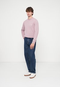 Camisola de manga comprida rosa clara com um pequeno logótipo, combinada com jeans azuis folgados e sapatilhas brancas com solas marrons.