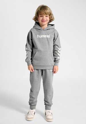 Smilende barn med krøllet blondt hår, der står, iklædt en grå hummel hættetrøje, matchende joggingbukser og hvide sneakers med sorte striber.