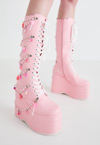Bottes plateformes roses jusqu'au genou en matériau lisse, dotées de lacets blancs, de ferrures en argent et de chaînes décoratives avec des charms colorés.