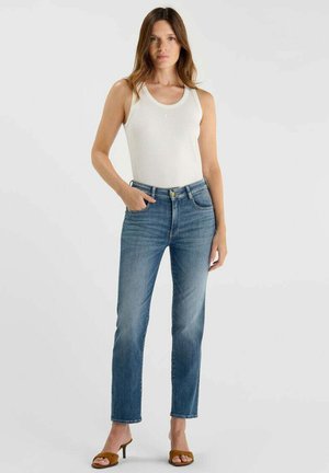 Jeune femme portant un haut blanc sans manches, un jean bleu droit et des sandales à talons marron, debout avec une main dans la poche.