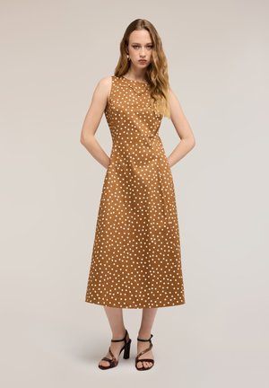 Donna che indossa un vestito midi marrone senza maniche con pois bianchi e sandali neri con tacco alto, in piedi con le mani dietro la schiena.