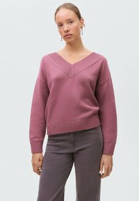 Pulover z V-izrezom v pastelno roza barvi z rebrastim izrezom in manšetami, ki ima sproščen kroj in teksturiran pleten vzorec. Ujemajo se s sivo hlačami.