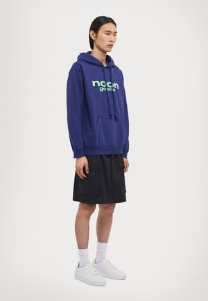 Granatowy hoodie z zielonym logo, w połączeniu z czarnymi szortami i białymi sneakerami. Posiada kieszeń kangurkę oraz kaptur ze sznurkiem.