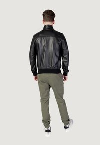 Giubbotto bomber in pelle nera con polsini e orlo a coste, indossato sopra pantaloni verde oliva. Presenta un colletto alto e una texture liscia.