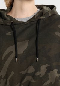 Camo-patterned hoodie in dark green and black with a drawstring hood, silver metal accents, and a soft texture. Layers with a gray collar visible.