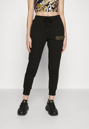 Femme portant un jogging noir Versace Jeans Couture avec une taille à cordon et des baskets noires assorties avec des logos dorés.