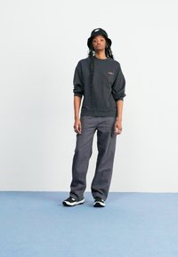 Mørk grå oversized sweatshirt med brodert logo, kombinert med løse grå bukser og svart-hvite sneakers. En svart bøttehatt fullfører antrekket.